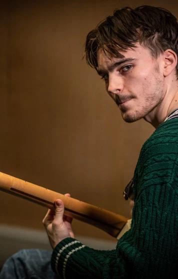 A young guitarist wearing a green sweater, holding a Fender guitar, and looking towards the camera in a dimly lit practice room. A young guitarist wearing a green sweater, holding a Fender guitar, and looking towards the camera in a dimly lit practice room.